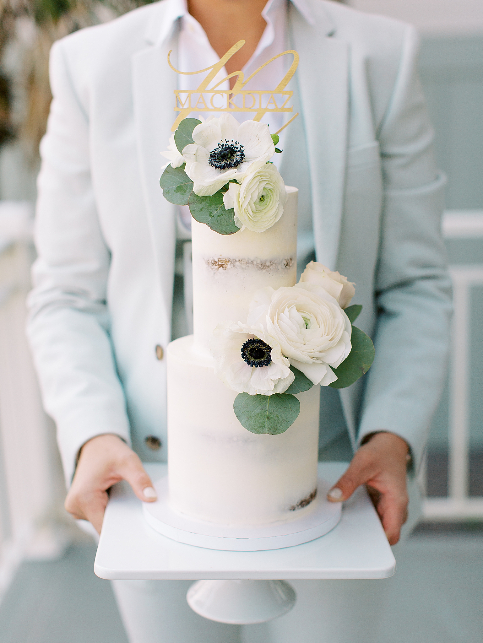 bride in blue suit holds naked wedding cake with floral accents