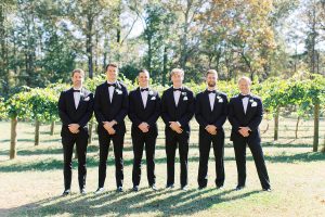 groom and groomsmen in classic black tuxes pose along farm