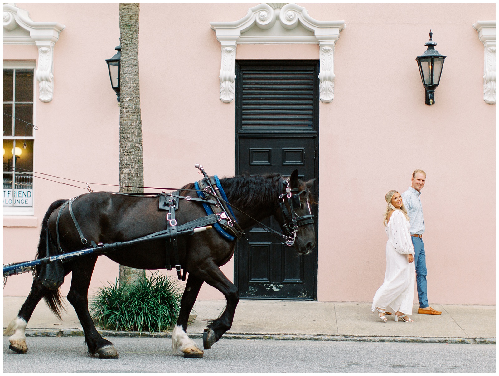 couple walks along pink house in Charleston with carriage behind them