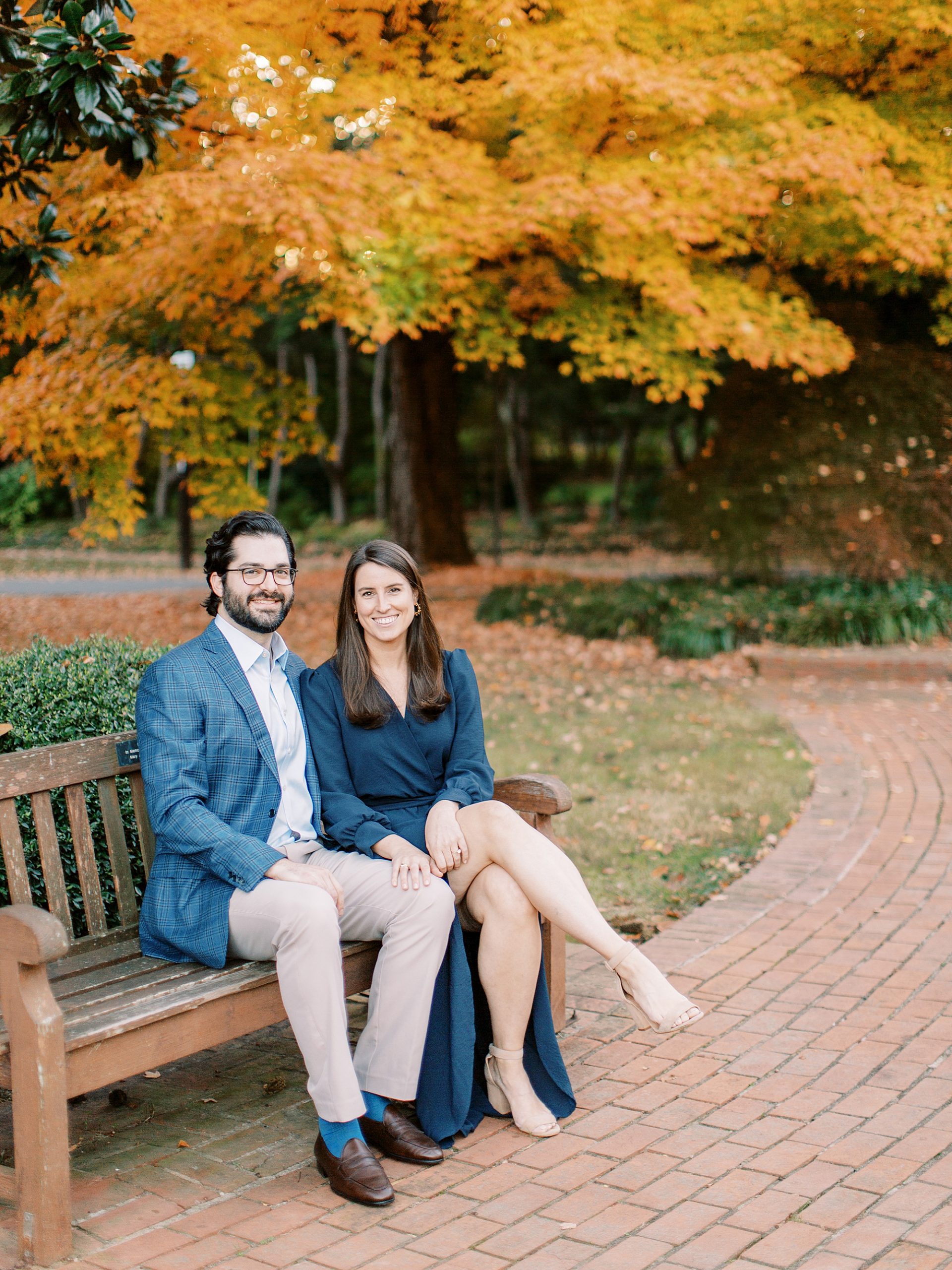 engaged couple sits on wooden bench during fall engagement portraits