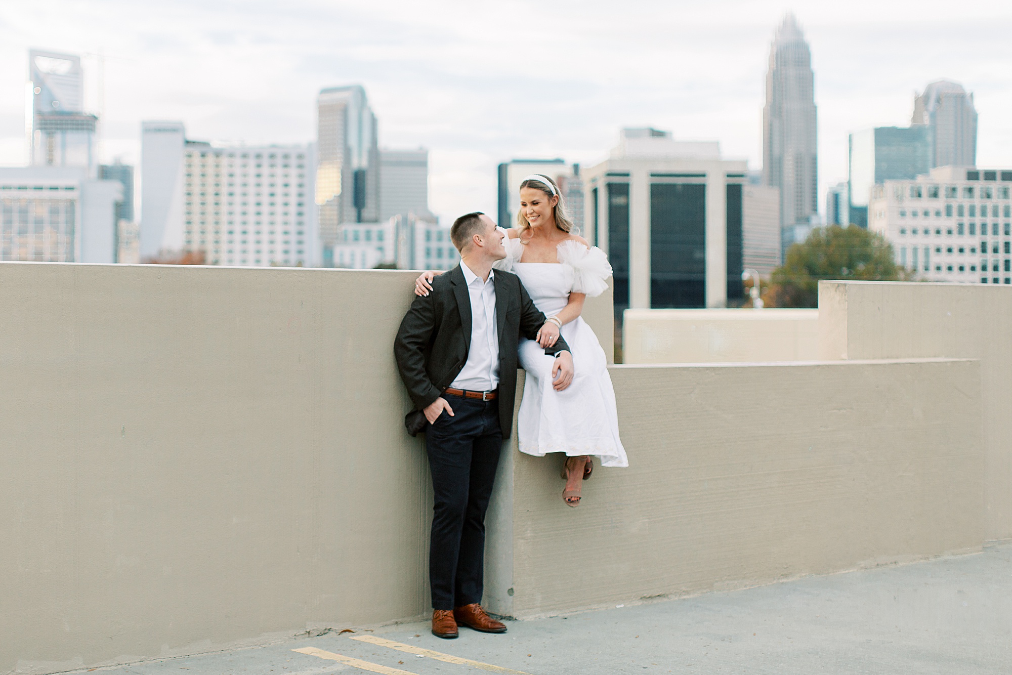 bride sits on wall with Charlotte NC skyline behind her