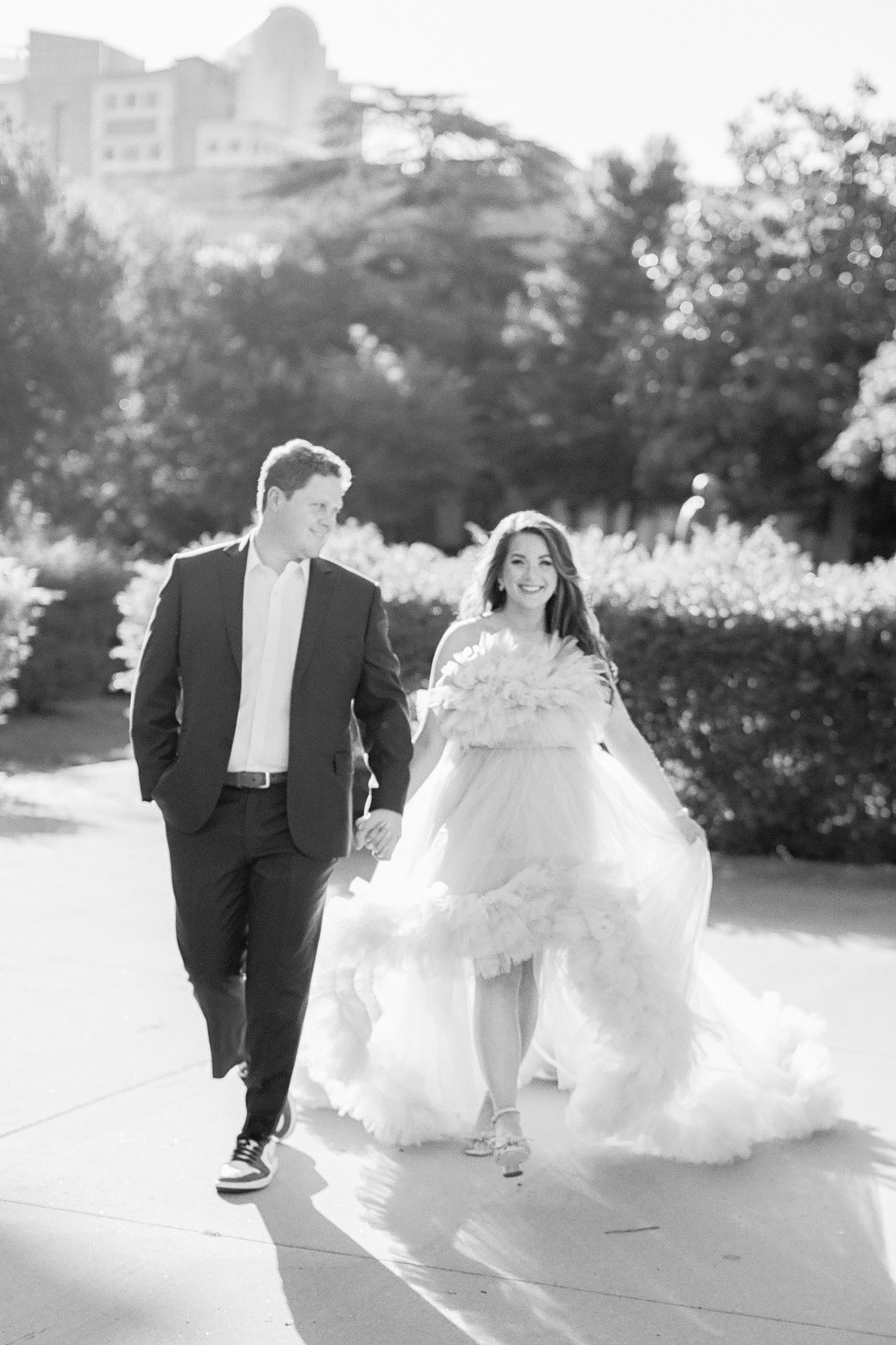 couple holds hands walking through Charlotte park with bride in dramatic gown