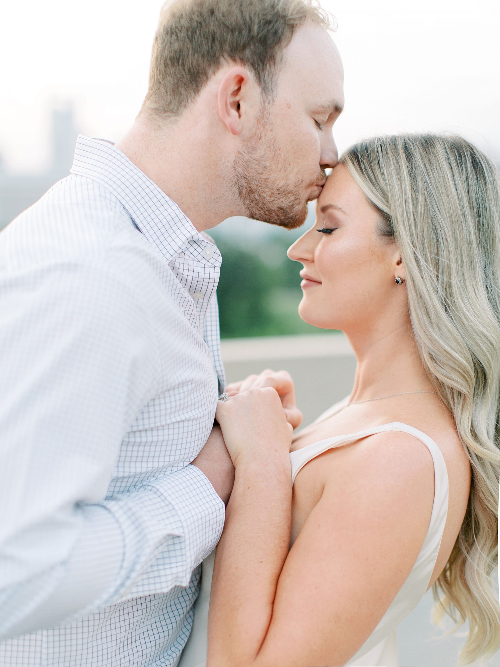 bride and groom hold hands while he kisses her forehead during Uptown Charlotte engagement session