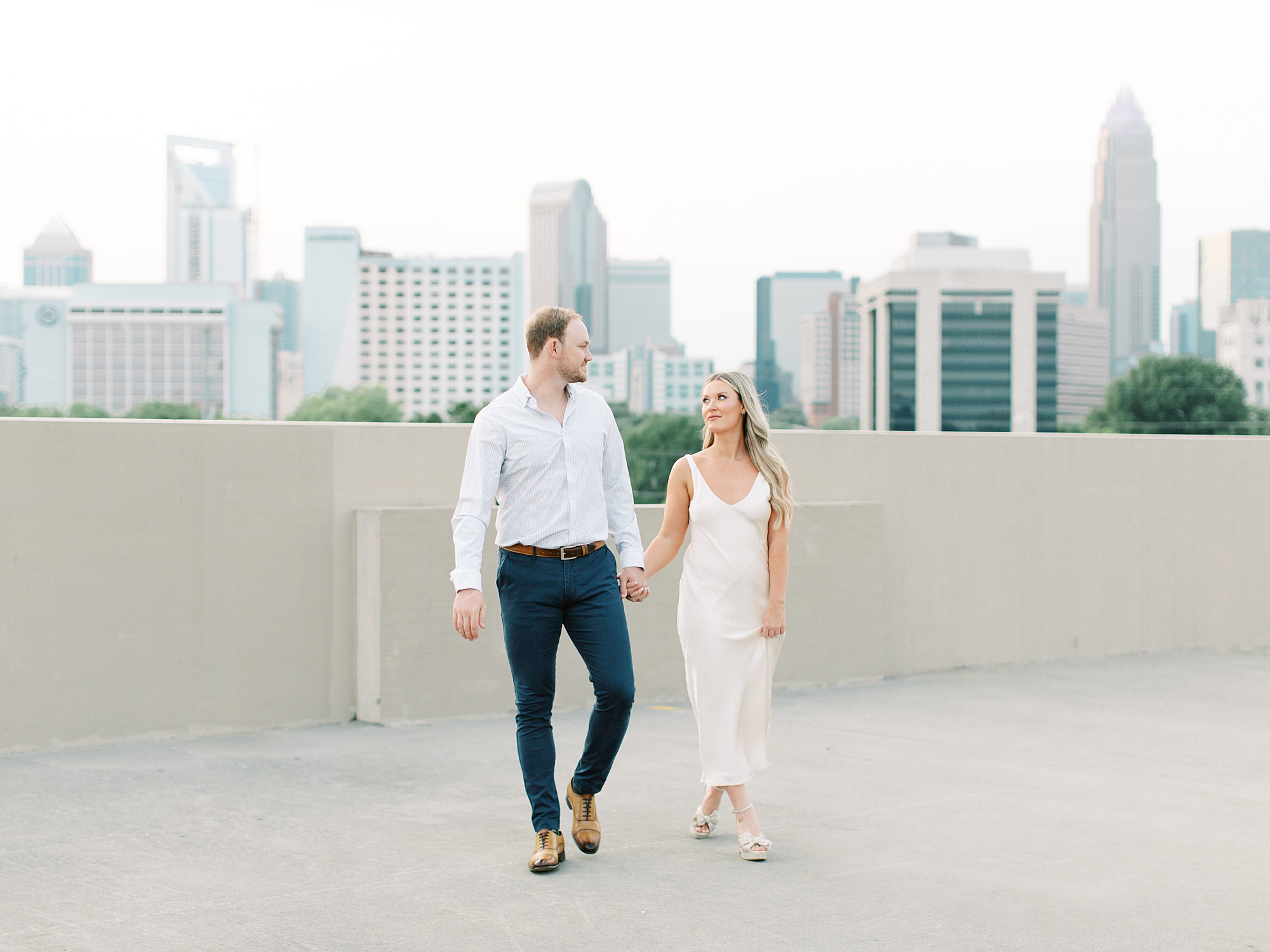 woman in white dress holds hands with fiancee walking on rooftop during Uptown Charlotte engagement session