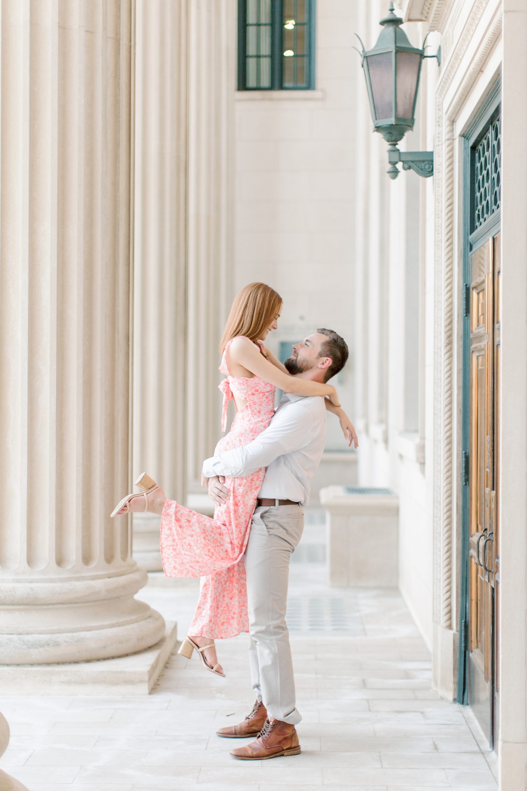 groom lifts bride in pink jumpsuit during Uptown Charlotte engagement photos