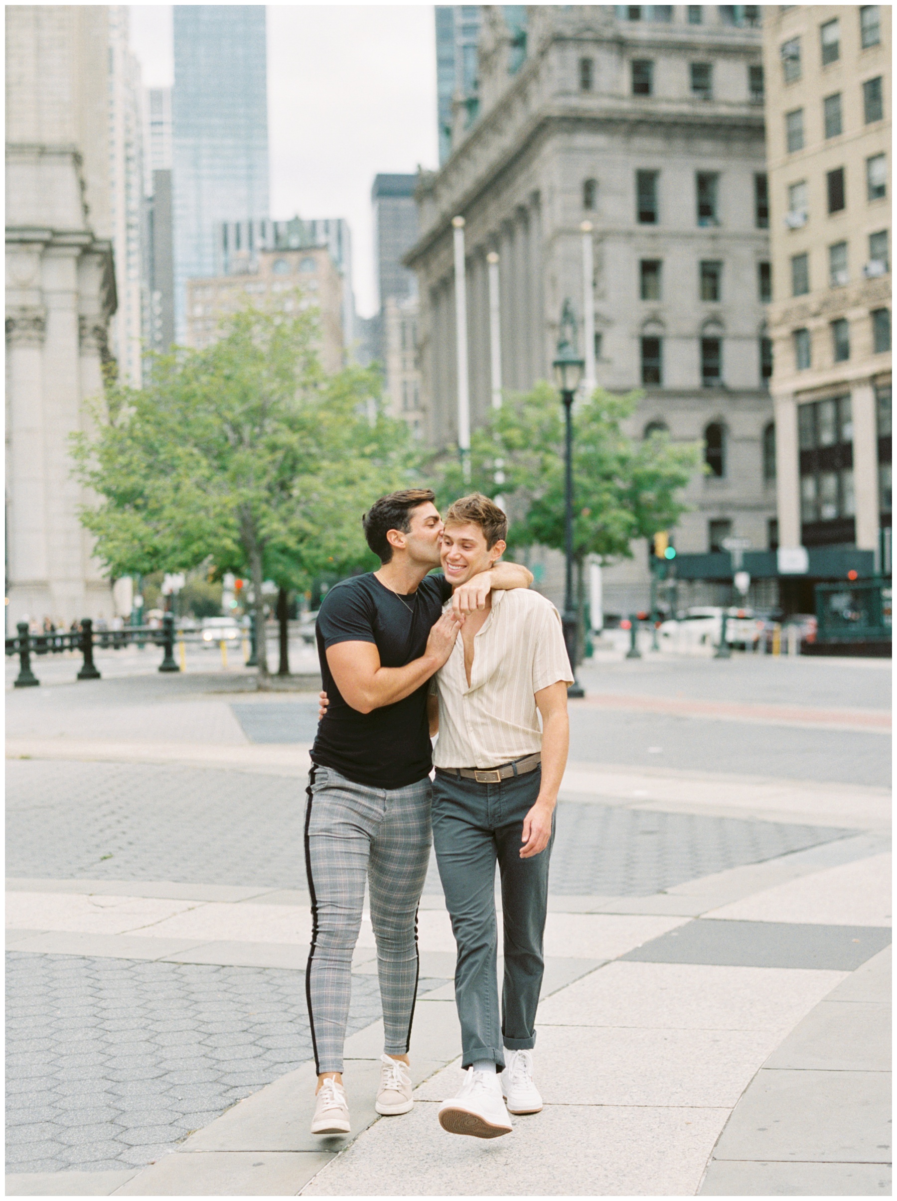 man hugs boyfriend around the neck kissing his cheek in Foley Square in NYC