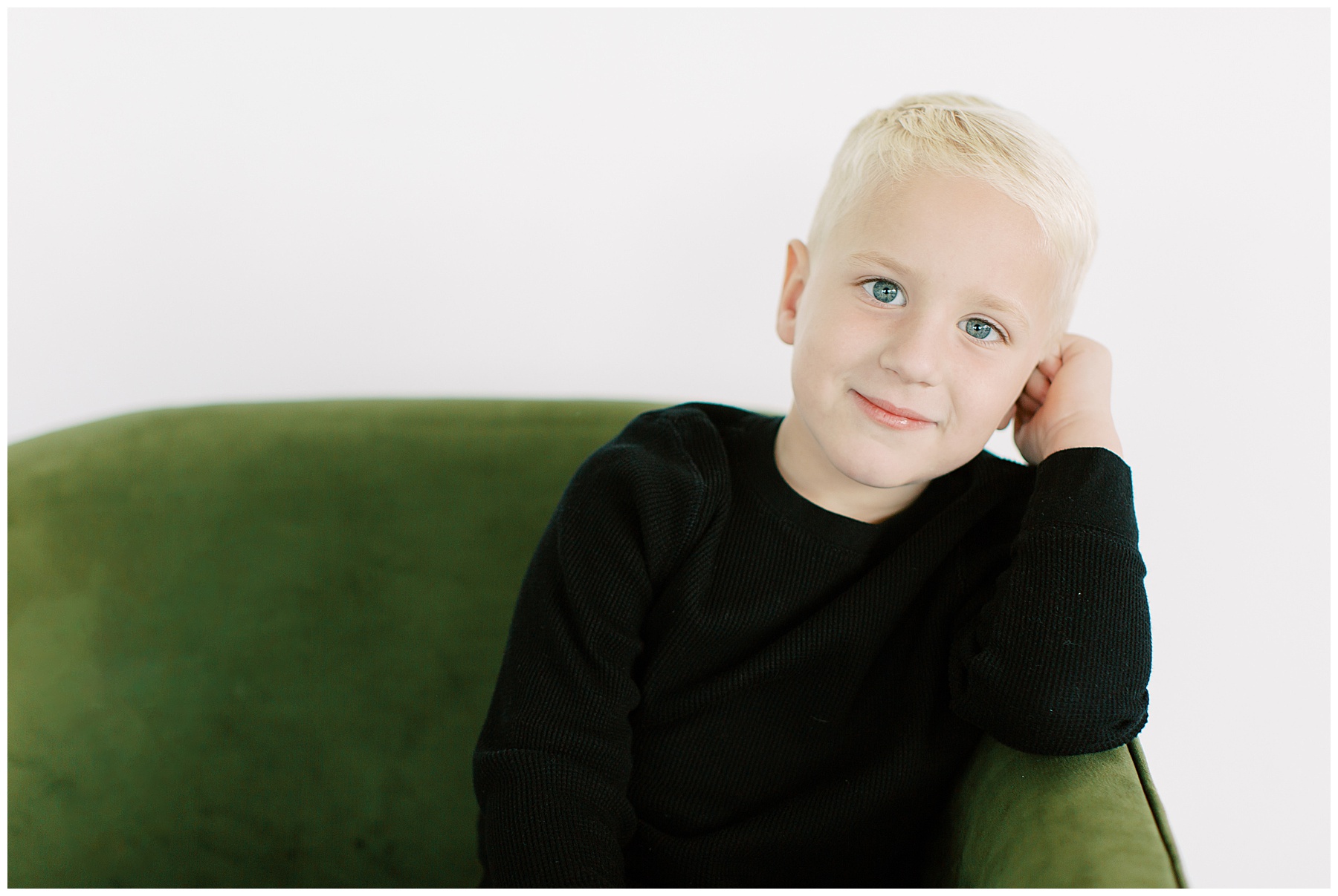 blonde boy with blue eyes leans on hand sitting on velvet green sofa during studio family photos 