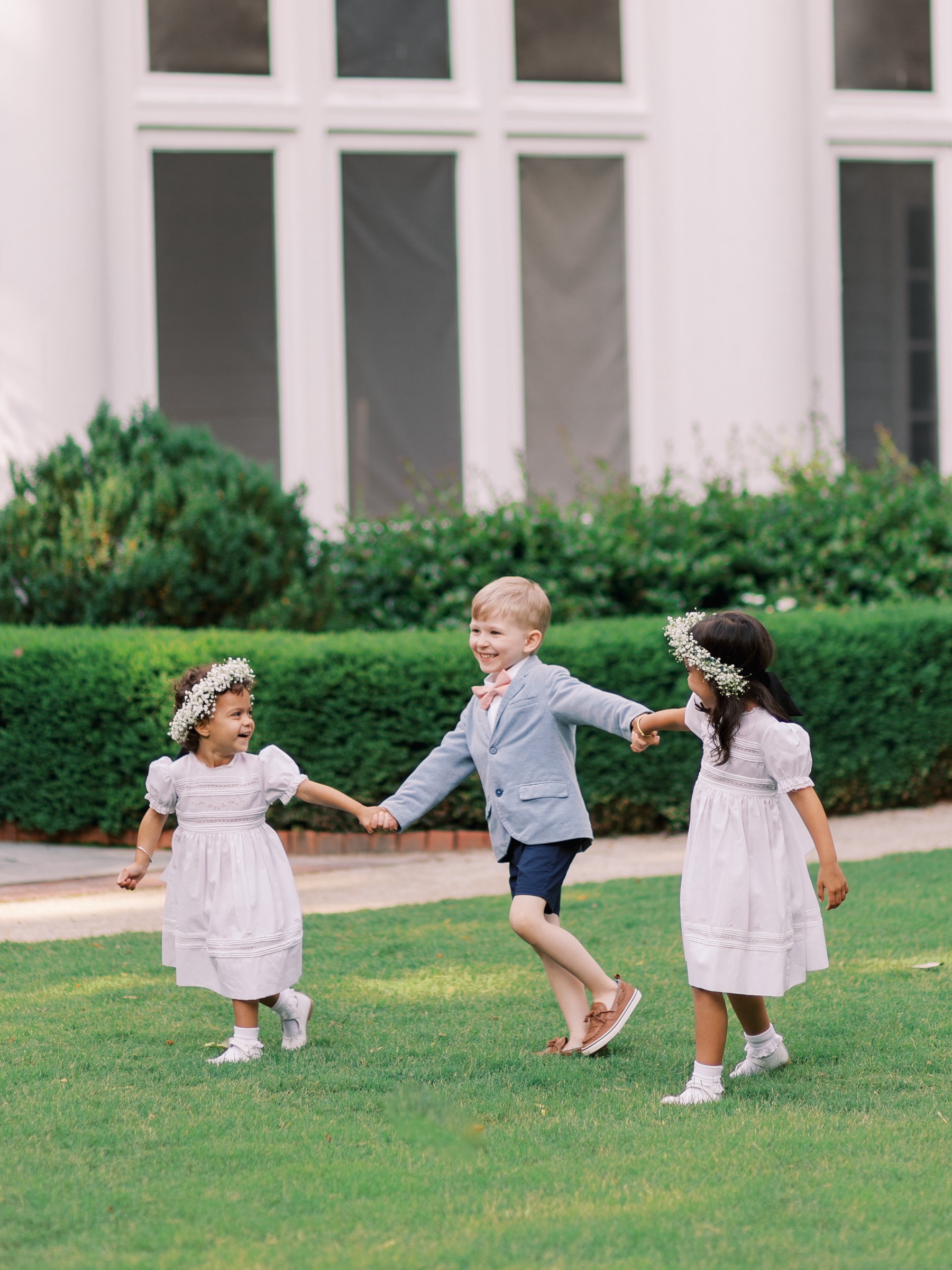 ring bearer and two flower girls hold hands walking on lawn at Duke Mansion 