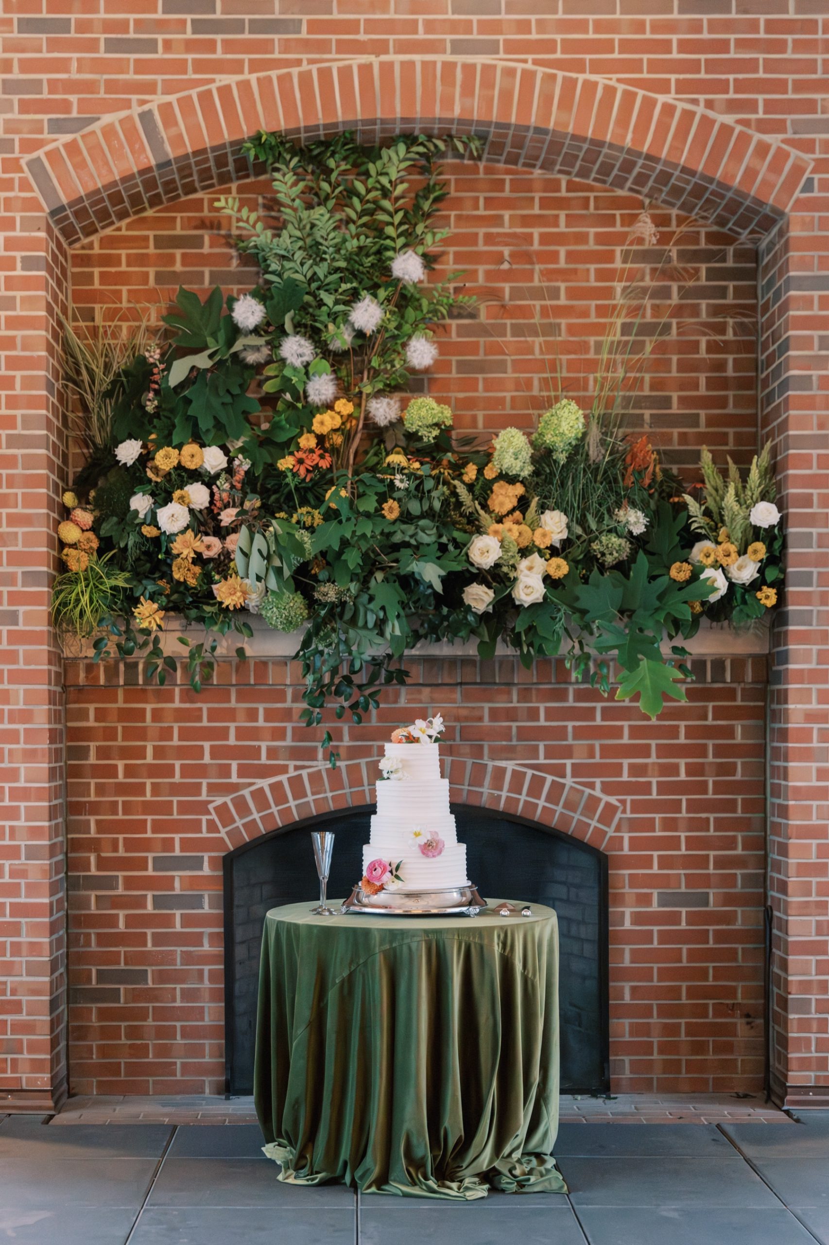 Wedding cake surrounded by flowers on the nearby fireplace mantle 